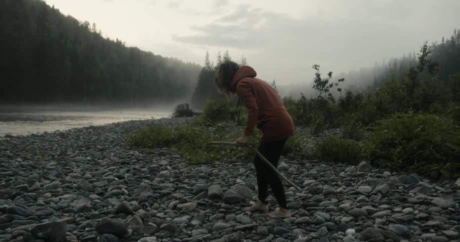 A woman walks across a stone pathway to a river and picks up a long stick