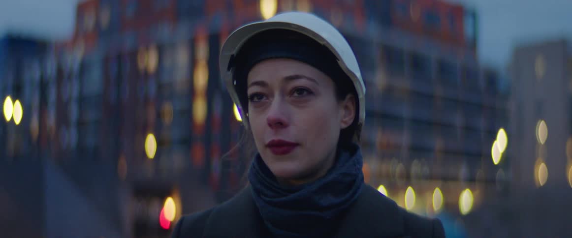 White woman wearing winter clothing and a white safety helmet talking to someone at a construction site in the middle of a big city