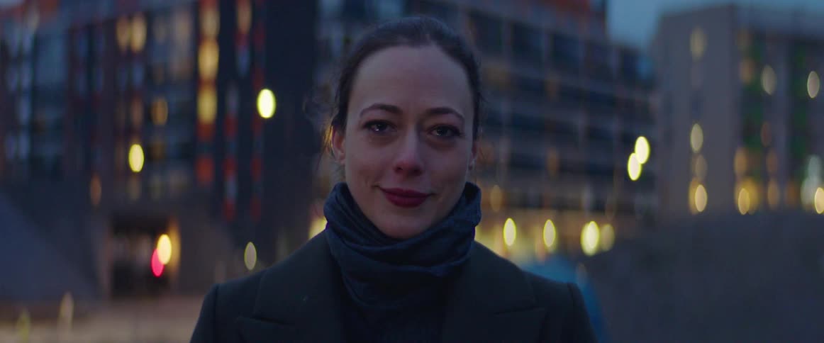 Woman with scarf and black coat faces the camera and cries tears of joy in the street with tall buildings in the background at dusk