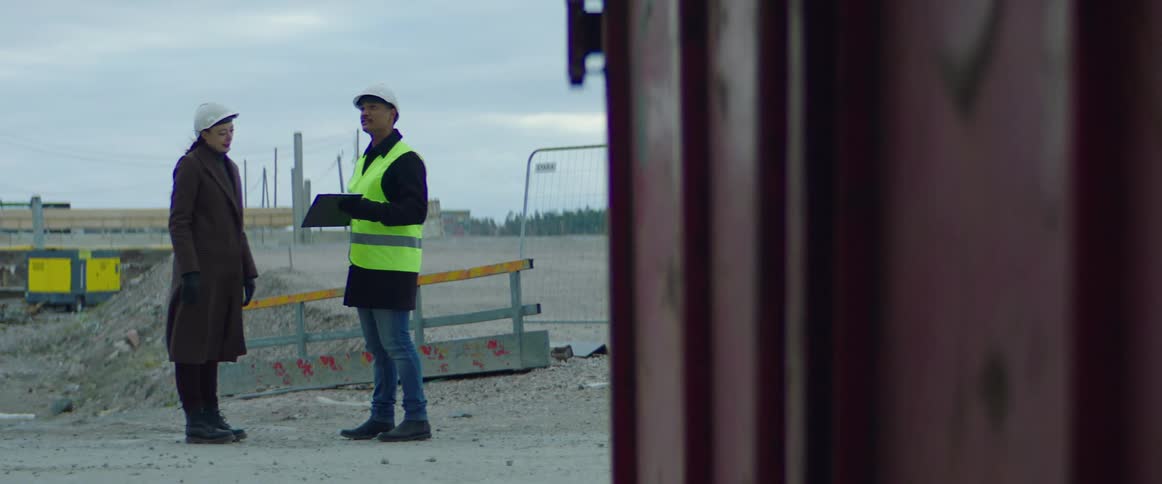 White woman wearing a safety helmet and overcoat shaking the hand and talking to a man holding a clipboard and wearing a green vest and safety helmet at a construction site