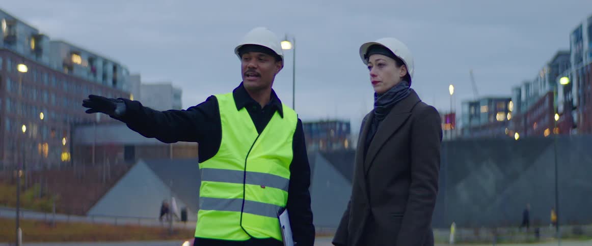 A construction worker wearing a vest and a hardhat meets a woman wearing a hardhat at a construction site and they shake hands and talk