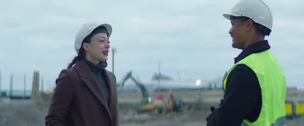 A male construction worker holding a clipboard and wearing a safety helmet and green vest talking to a woman wearing a safety helmet in the middle of a construction site