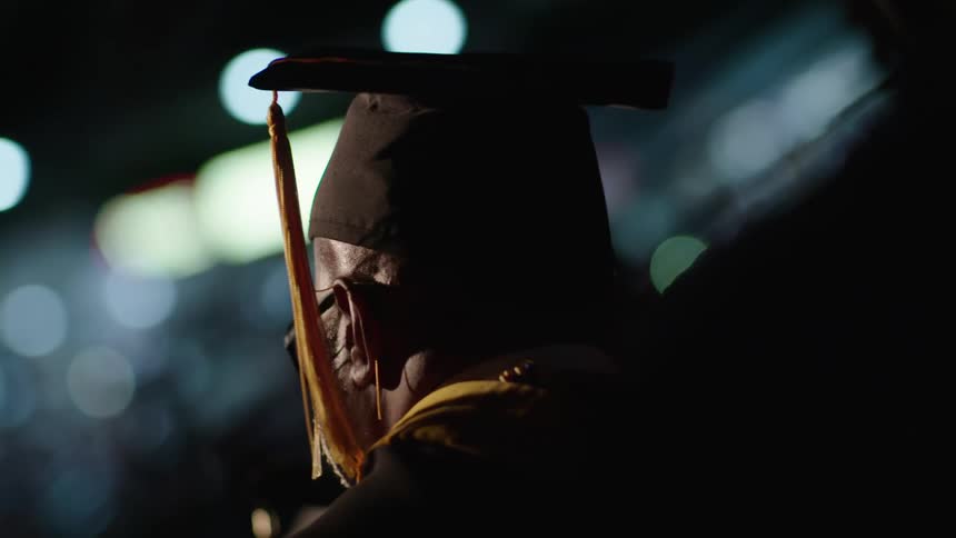 A black man wearing a graduation cap stands at a ceremony