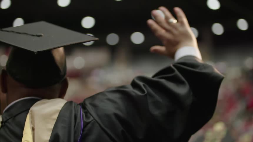 Black man wearing graduation hat and graduation robe celebrating and waving at the crowd during a graduation ceremony