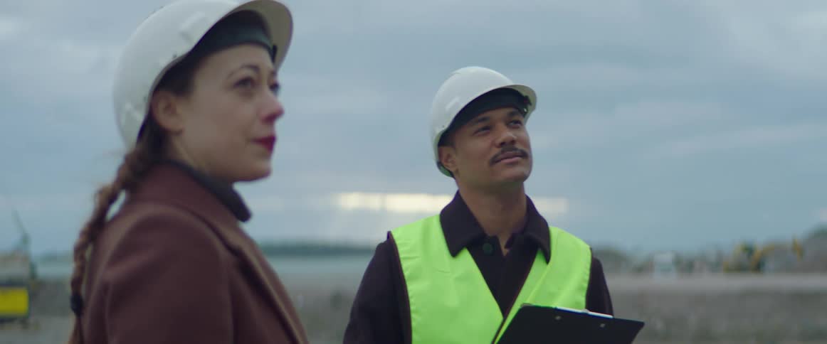 Man holding a clipboard and wearing a safety helmet and green vest shaking the hand and talking to a woman wearing a overcoat and a safety helmet at a construction site outdoors