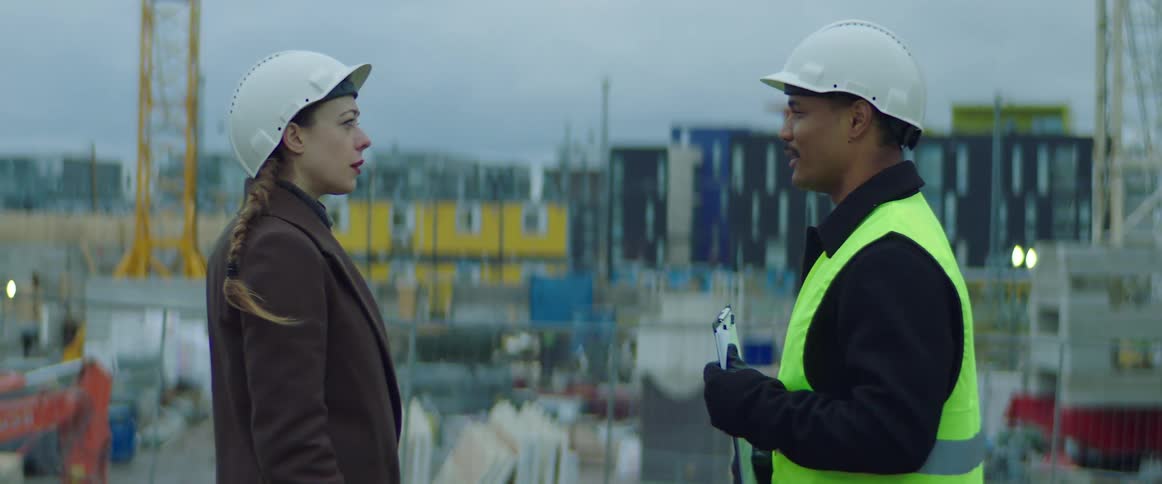 Two coworkers stand at construction site taking while one of them holds a clipboard
