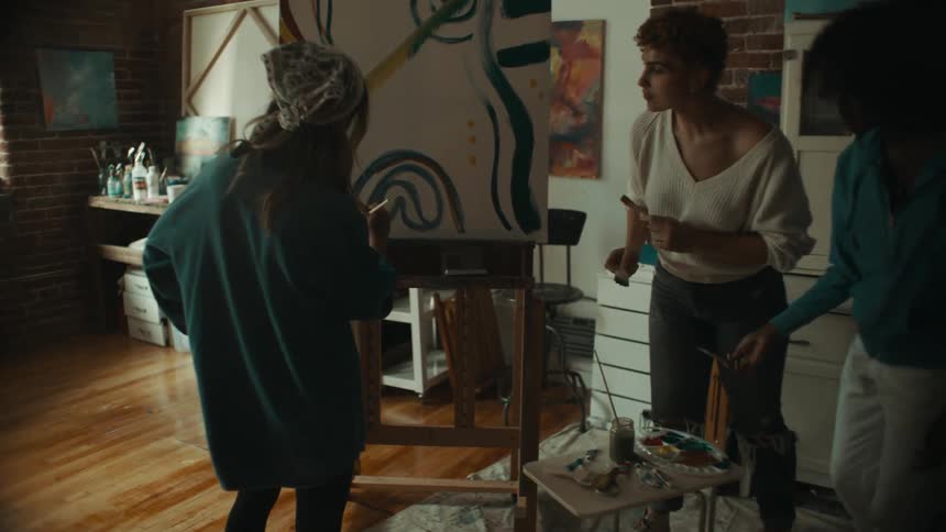 Three women paint on the artwork that is sitting on an easel in a large room