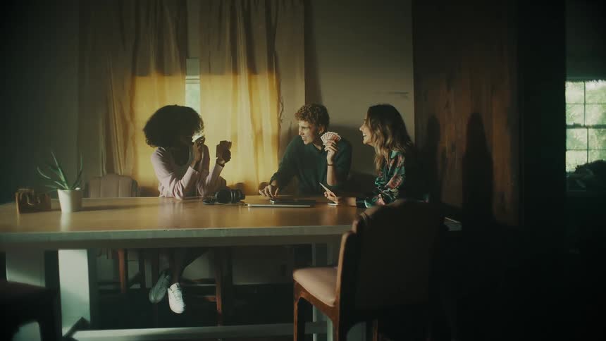 Three teenagers talk and laugh as they play cards inside a house