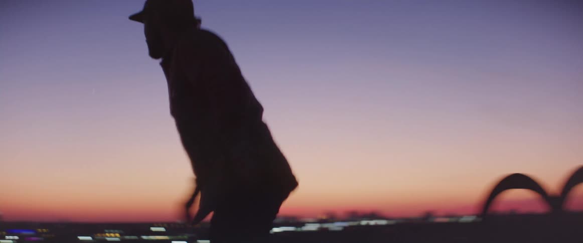 Man climbing an access ladder to the building rooftop and running to the edge of it during sunset with a big city in the background