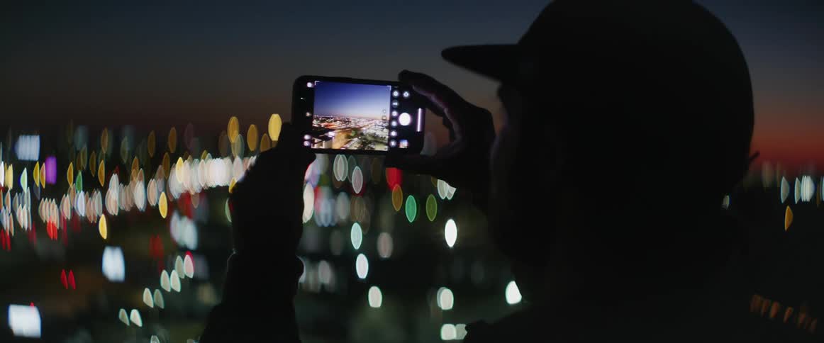 Man stands taking pictures of city lights spreading toward red sky on the horizon