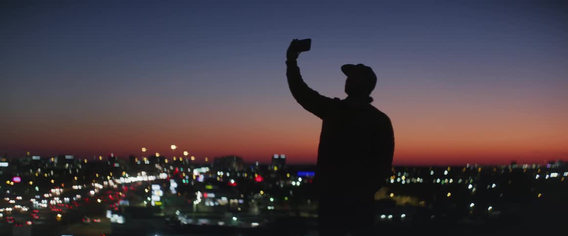 A man takes a selfie with his smartphone while standing on a hill with views of a bustling city