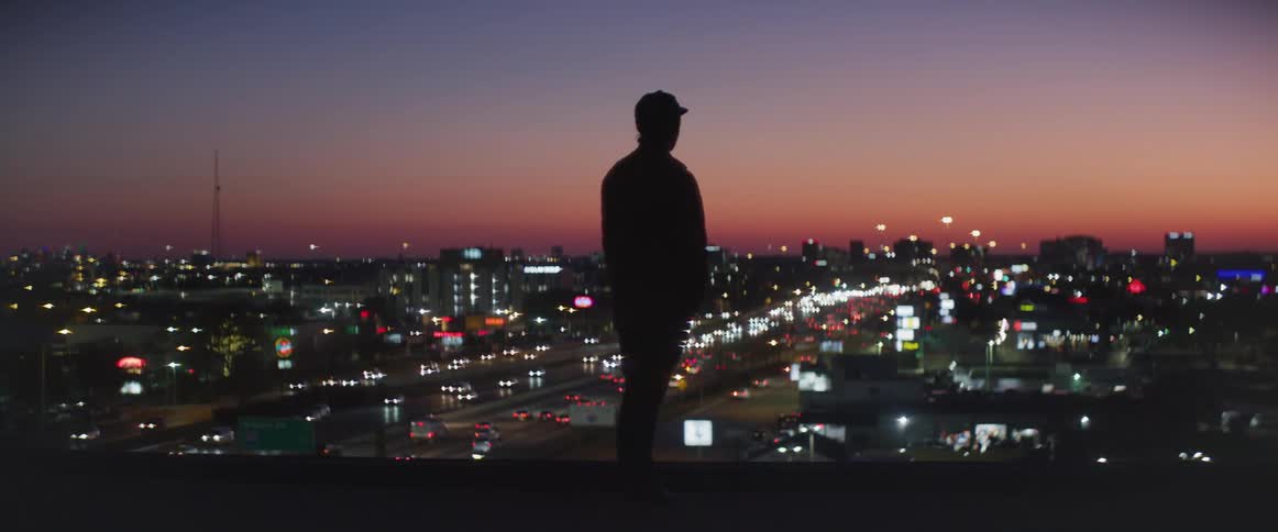 A man is standing on top of a building overlooking a city and freeway traffic at night