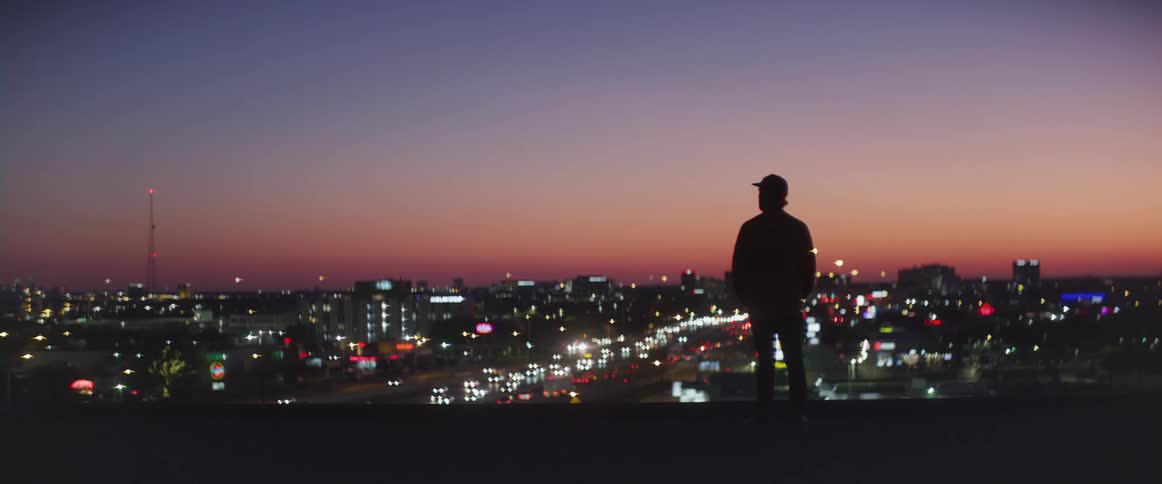 Man stands silhouetted against red horizon watching heavy traffic on highway through the city at night