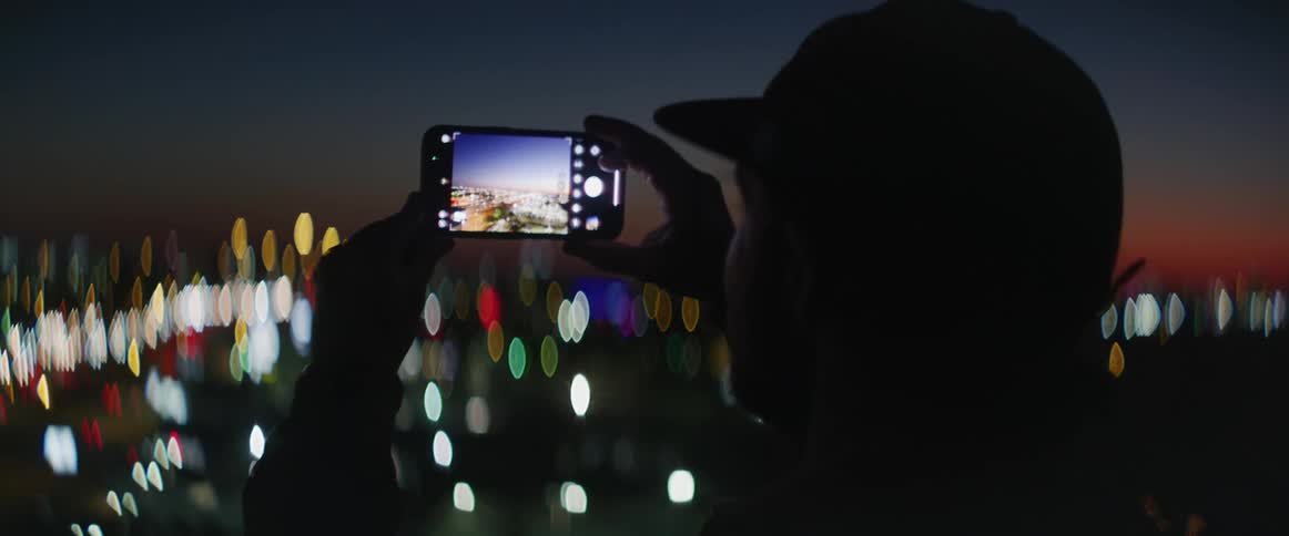 A man holds up his smartphone and snaps pictures of the city and traffic below at night