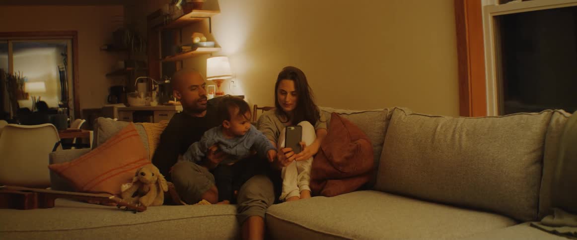 A woman sitting on the couch next to her husband holds a cell phone and laughs as he lifts their son above his head