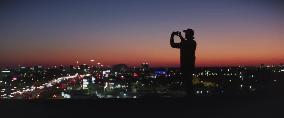 Man standing at the top of a building taking pictures of the city ahead during sunset