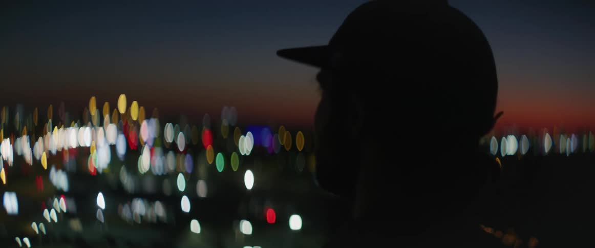 A man holds up his phone and snaps a photo of the city below him with lights glowing