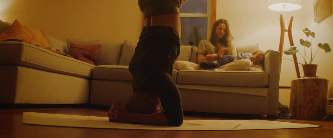 A person practicing yoga in a yoga mat at the living room while a teenager girl and a toddler are resting at the couch