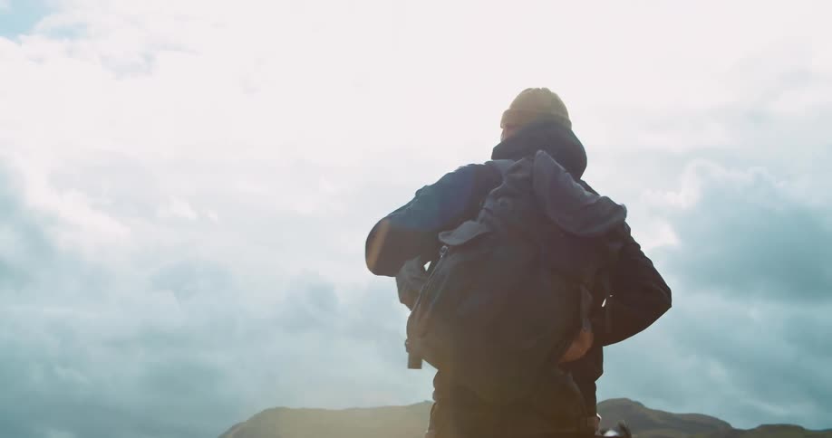 A man bends down and ties his backpack and then stands up and puts it on and walks down a hill