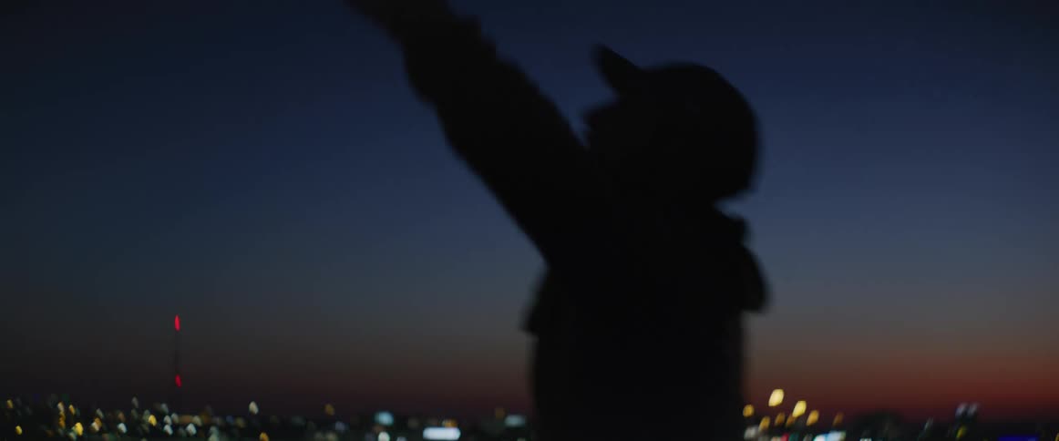 Man silhouetted against red horizon above brightly lit city at night spins with his arms outstretched