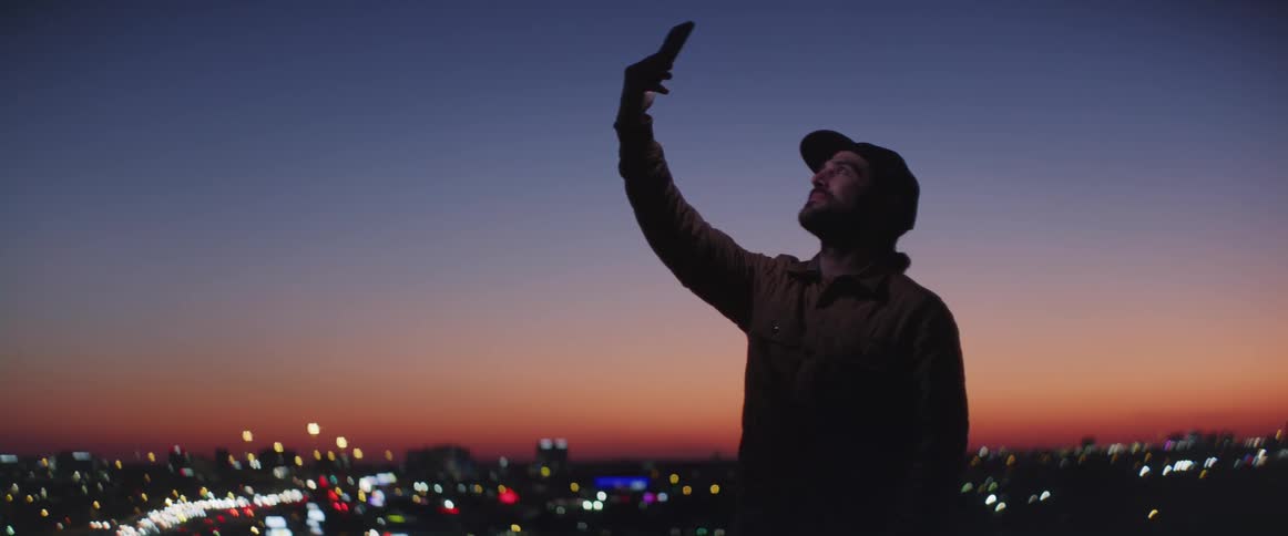 Man stands outdoors scanning the sky with a smartphone with city lights stretching to the red horizon at night