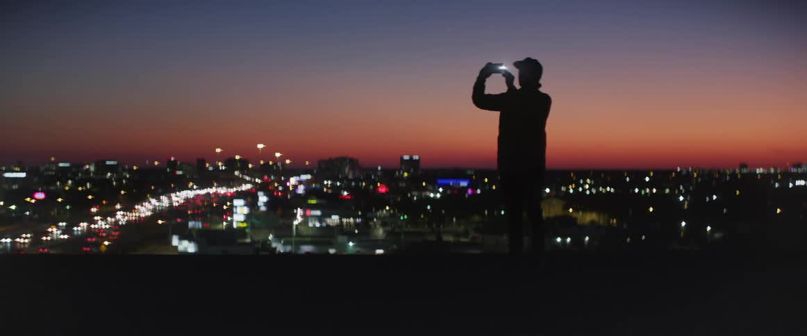 Man standing in the top of a building taking selfies with his smartphone with a view of a big city behind him during sunset