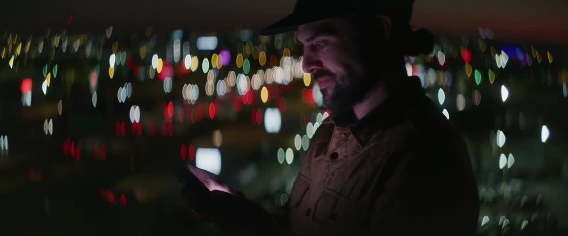 White man wearing a baseball cap standing in the top of a building tapping and scrolling his smartphone