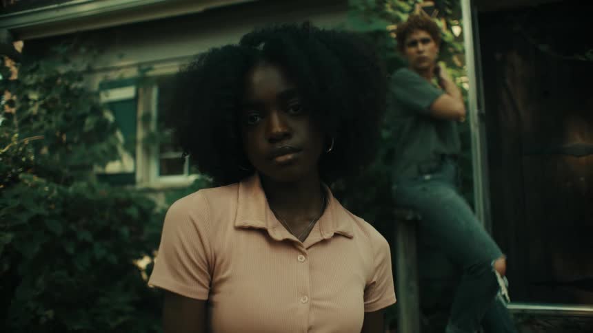 A teenage girl leans on a railing outside a house as another girl stands in front of her