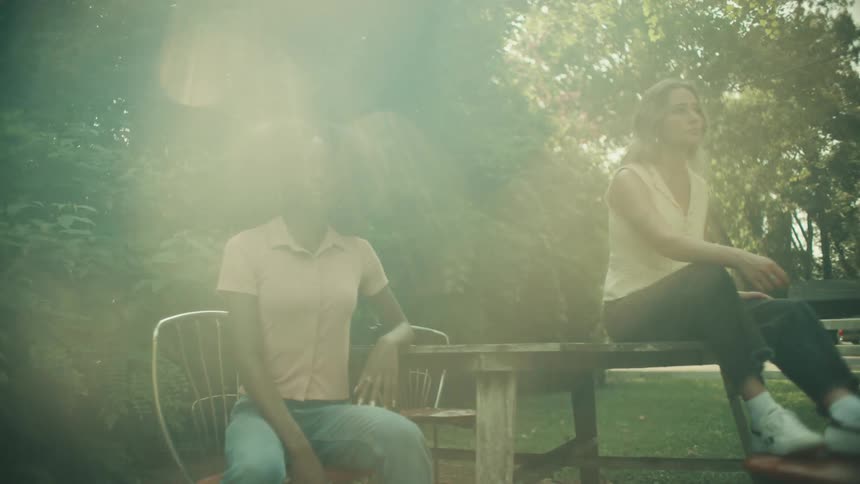 Black woman sitting in a chair next to a wooden table while two other women are sitting next to her at the table and at the chair