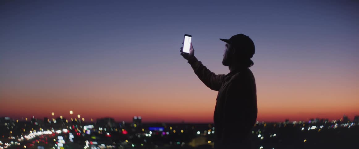White man wearing a baseball cap holding his smartphone and video calling someone with a big city behind him during sunset