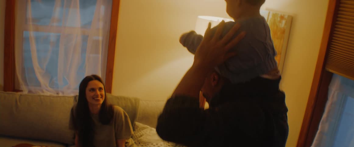 A woman sits on a couch watching a man bounce a baby on his shoulders