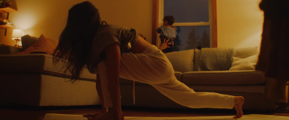 Woman does yoga near the sofa while a man and baby look out the window