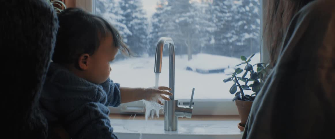 A woman stands next to her husband at the kitchen sink as he holds their baby who runs his hand under the running faucet