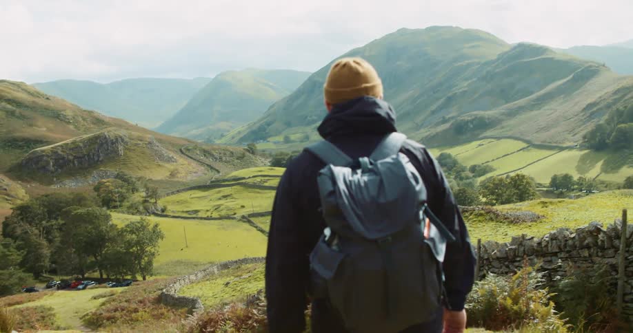 Man wears a toque and has a backpack on his back and walks towards a green field between the hills