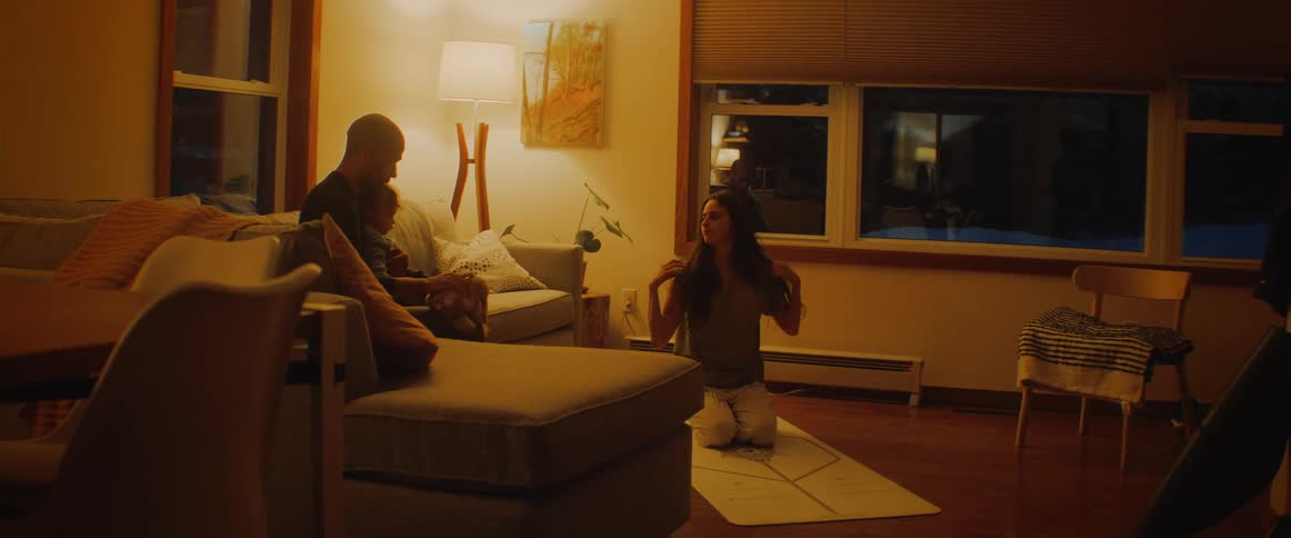 Woman doing yoga at a yoga matin the living room while her husband and baby son are sitting in the couch playing with a plush