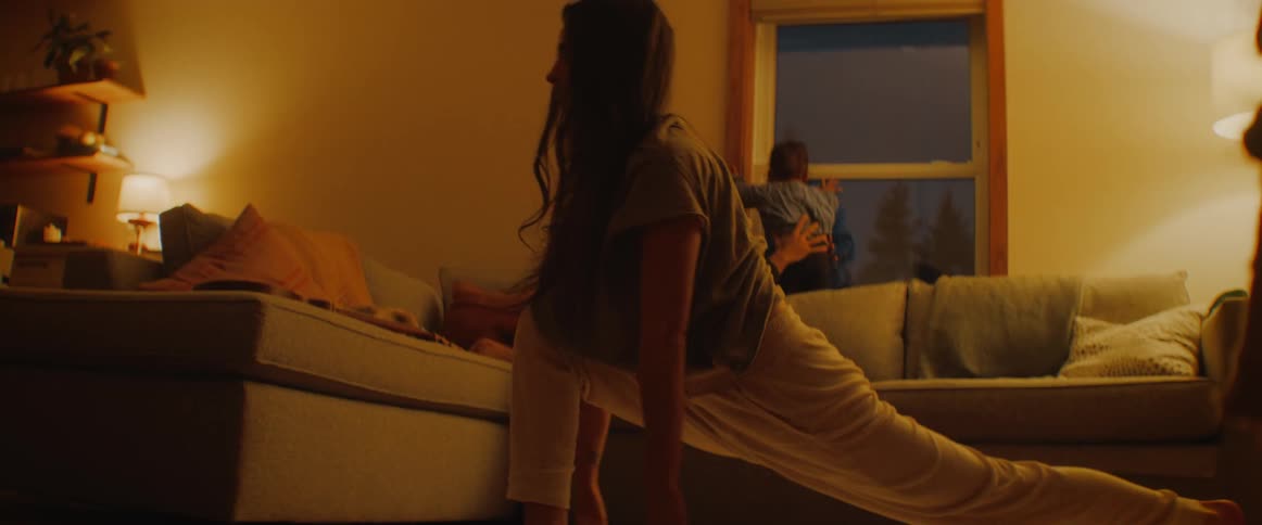 Family in the living room while father and son are sitting in the couch next to a window and the mother is doing yoga in the floor