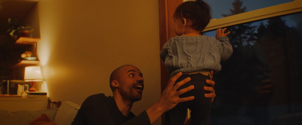 A man holds his young son as he tries to stand and look out a window at home