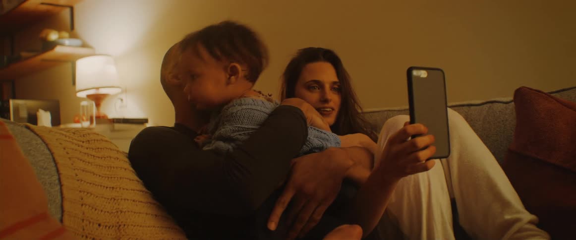 A woman hold a cell phone having a video call with a baby crawling over a man and a couch