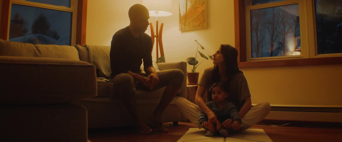 Mom sitting on a mat on the floor with baby in front of her while talking to dad on the couch