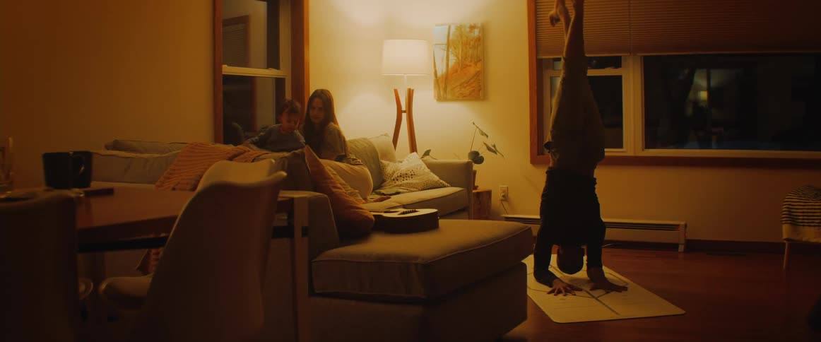 Father mother and son in the living room while the father is doing yoga at the yoga mat in the floor and the mother and baby are sitting in the couch looking through the window