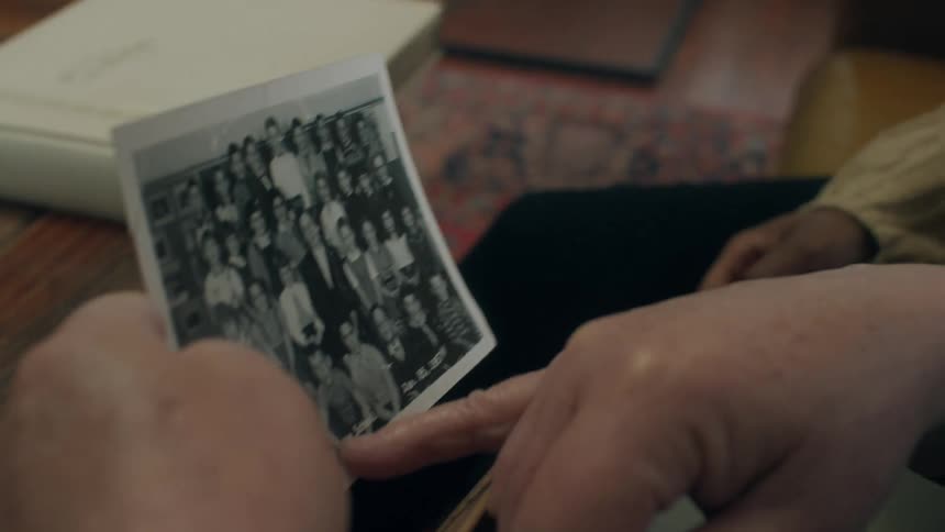 A young woman sits in a house with an elderly man and they look at an old school photo