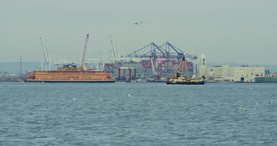 The staten island ferry moves away from shore with a plane flying overhead and cranes behind