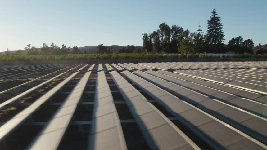 The sun is shining brightly on solar panels on a solar farm with trees and mountains in the distance