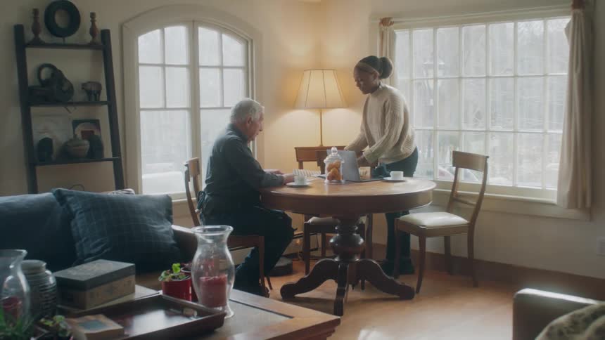 Black woman and old man talking and sitting at a table with coffee cups papers and a laptop on top