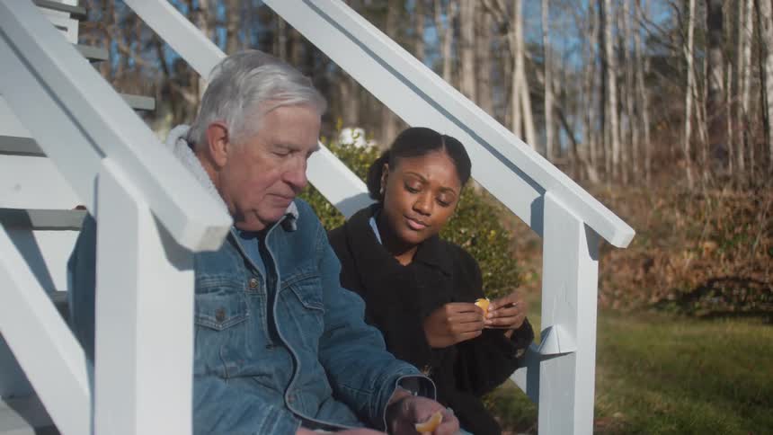 An elderly man sits next to a woman on stairs outside a home and they talk as they eat