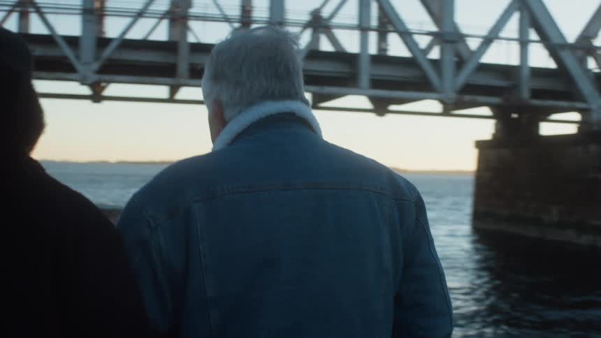 Woman walks arm in arm with elderly man along boardwalk passing bench