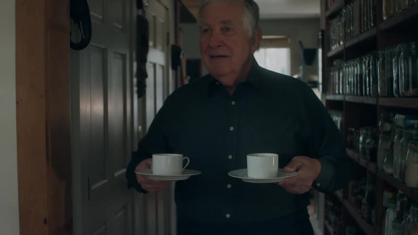 An elderly man walks down a hall in his home carrying two cups of coffee and lifts them up as he offers them