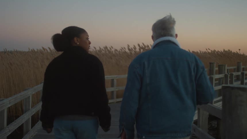 Elderly man and young woman walk on the pier over the water with reeds nearby