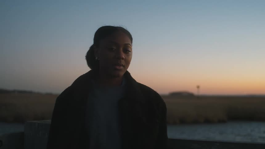 Woman with her hair in bun and black coat looks at camera near the lake at sunset