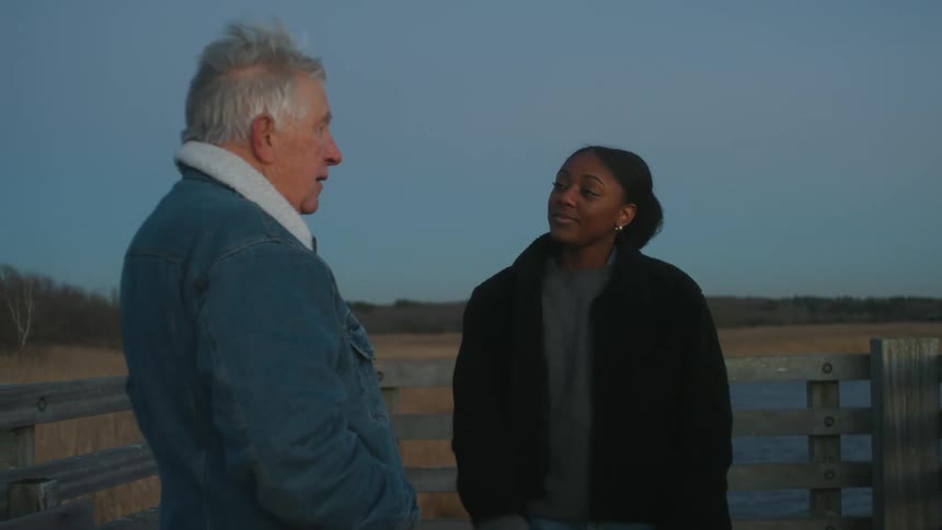 An elderly man and a young woman are standing on a pier near a lake and forest while having a conversation on a windy day at dusk
